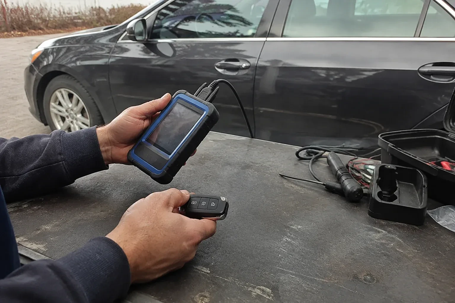 Hands holding a blue-edged electronic device and car-key fob at a worktable beside a parked car. — Transponder Key Programming, New Tecumseth
